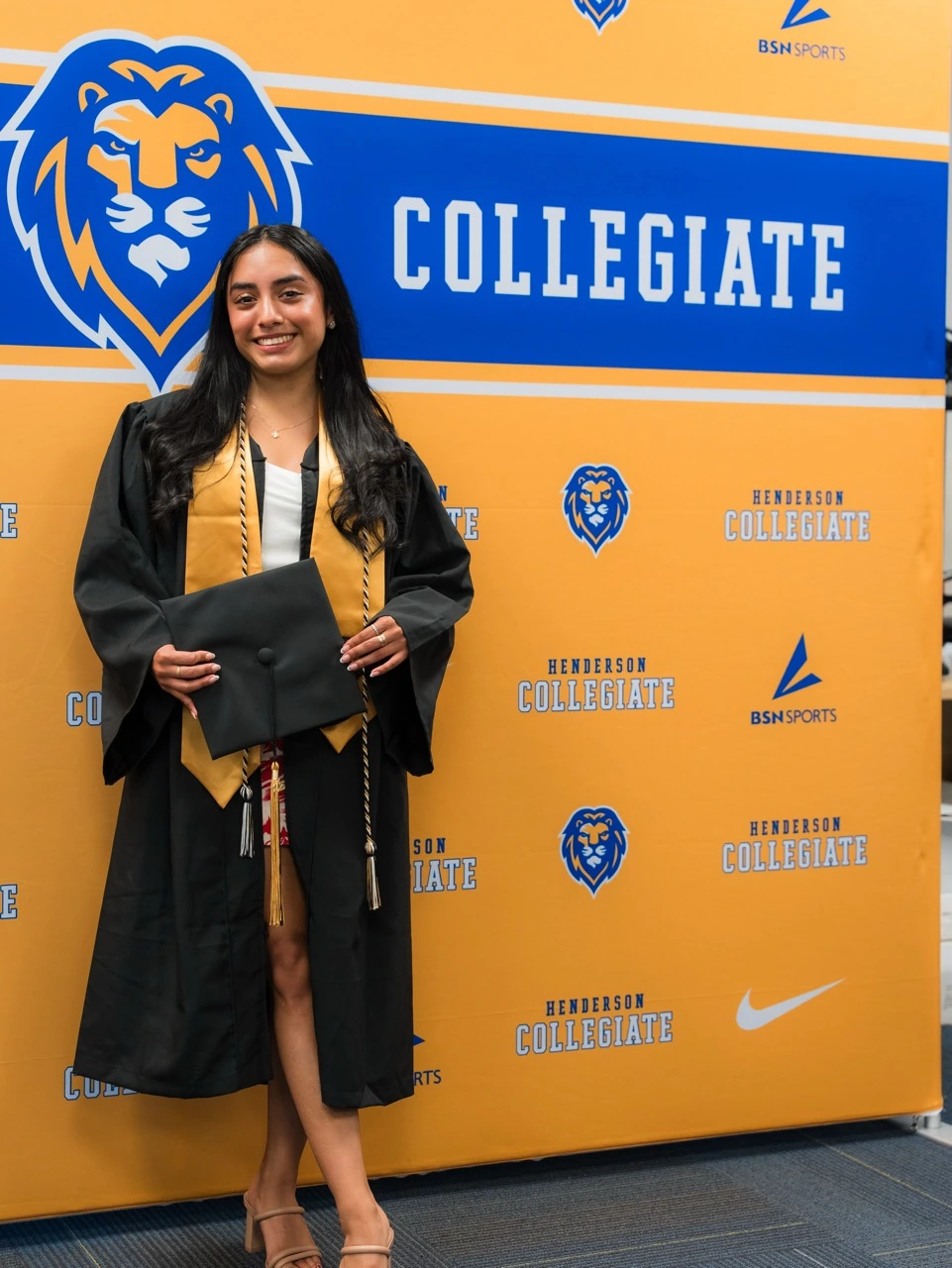 Graduate photo opportunity featuring student in black graduation gown holding diploma against Henderson Collegiate step-and-repeat banner with gold and blue background featuring repeated lion marks, wordmarks, Nike swoosh and BSN Sports logos for formal event photography