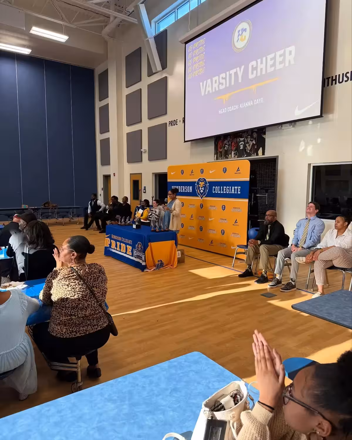 Varsity cheer assembly event in gymnasium featuring student athletes at branded table with Henderson Collegiate media backdrop, projection screen displaying 'VARSITY CHEER' announcement, demonstrating environmental applications for school athletic events and student recognition ceremonies