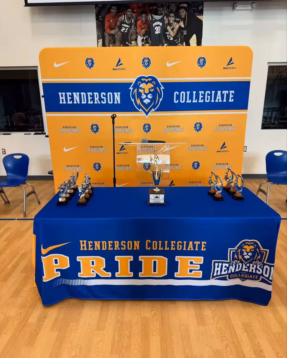 Trophy ceremony presentation featuring branded media backdrop with alternating gold and blue panels displaying Henderson Collegiate wordmark and lion marks, with championship trophies displayed on blue tablecloth with 'PRIDE' lettering and athletic achievements showcased