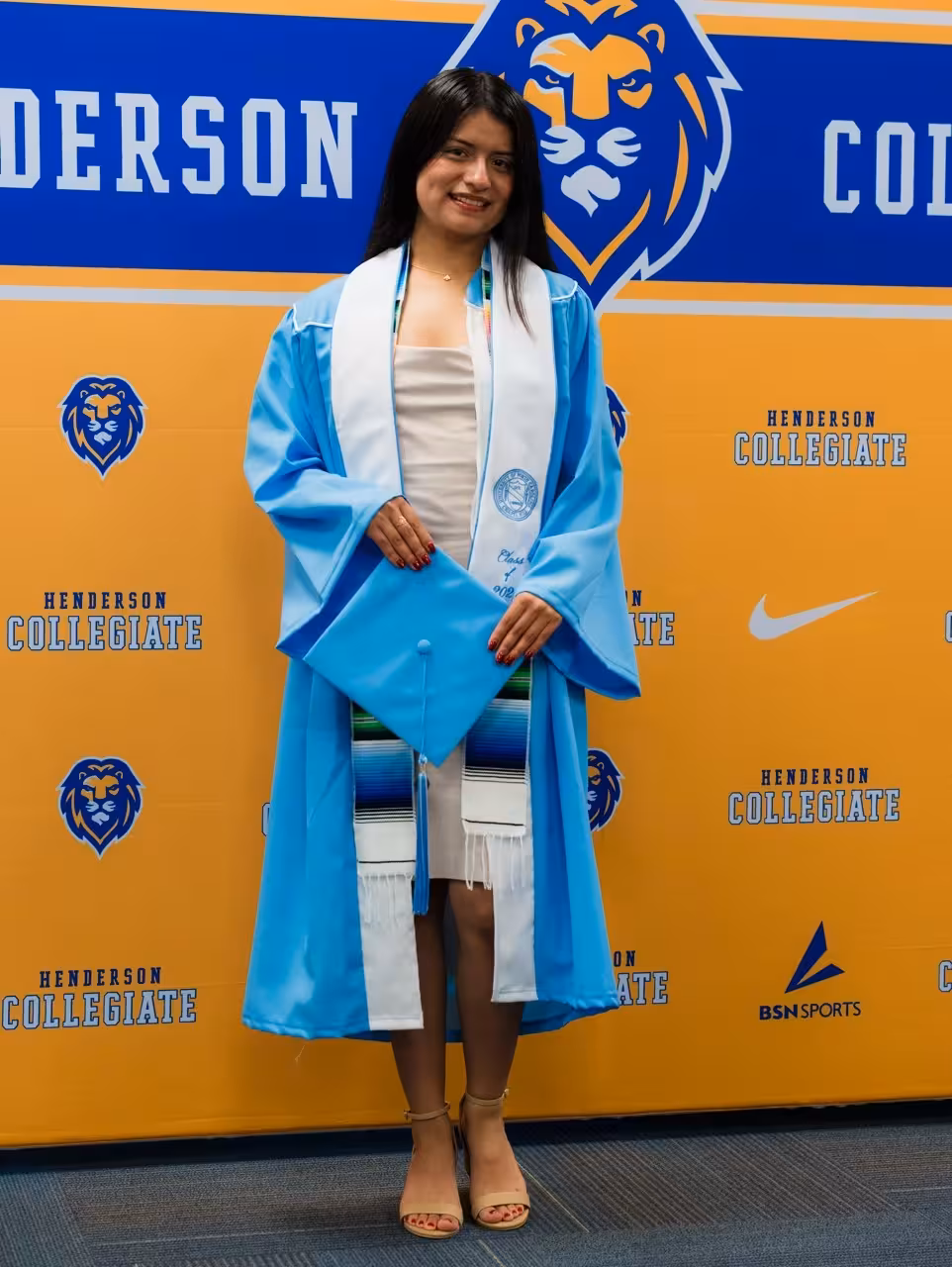 Graduate photo opportunity featuring student in light blue graduation gown against Henderson Collegiate step-and-repeat banner with gold and blue branded pattern, demonstrating versatile event backdrop system for commencement and academic milestone celebrations