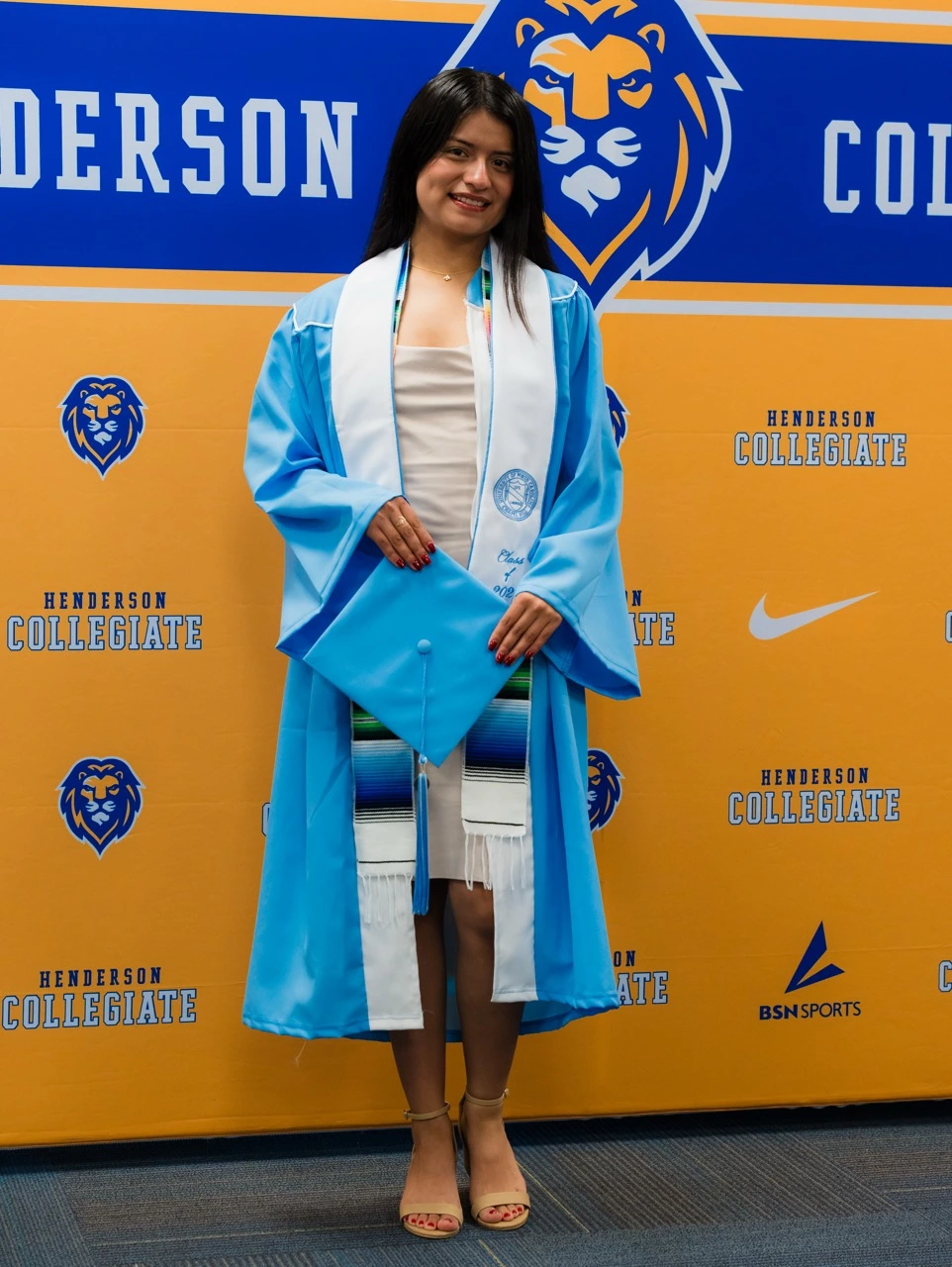 Graduate photo opportunity featuring student in light blue graduation gown against Henderson Collegiate step-and-repeat banner with gold and blue branded pattern, demonstrating versatile event backdrop system for commencement and academic milestone celebrations